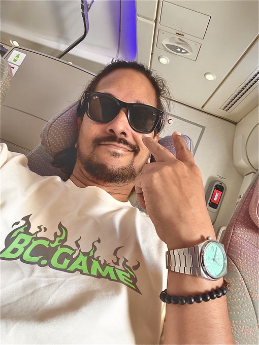 A man wearing sunglasses and a white "BC.GAME" t-shirt poses with a hand heart gesture while seated in what appears to be an airplane, sporting a turquoise watch and beaded bracelet. The image ties into his "Gm fam, happy sunday" post by projecting a relaxed, positive vibe—evoking a chill weekend vibe perfect for the crypto/gaming community associated with the BC.GAME brand.