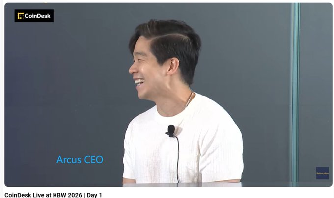 A still from a CoinDesk video showing an interview with the 'Arcus CEO' during 'CoinDesk Live at KBW 2026 | Day 1', where a smiling Asian man in a white shirt speaks into a microphone.