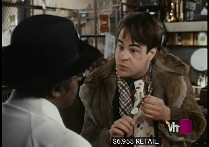 A VH1 TV show screenshot featuring a young man in a fur-collared jacket and patterned shirt holding merchandise, with a person in a black hat seen from behind; on-screen text reads '$6,955 RETAIL.'
