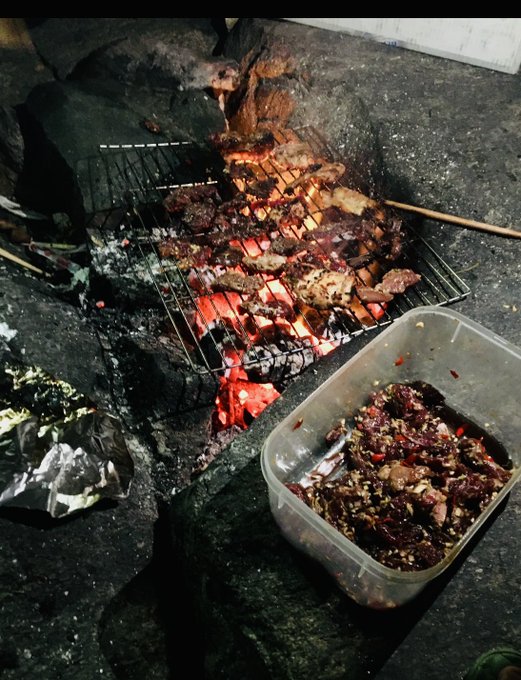 An outdoor nighttime scene showing meat being grilled over hot coals on a wire rack, with a plastic container of marinated raw meat beside it.