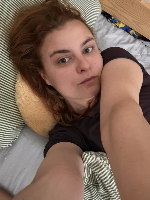 A selfie of a young woman with reddish-brown hair lying on a bed with striped pillows, taking the photo with her arm extended.