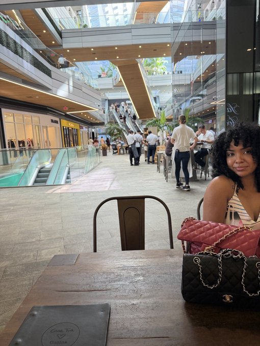 A woman with dark curly hair sits at a table with a black quilted Chanel handbag and a pink bag, located inside a modern, multi-level shopping mall with escalators and pedestrians in the background.