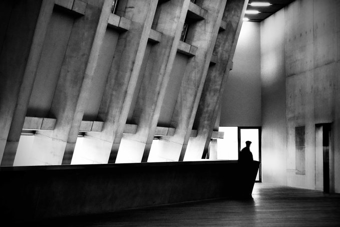 A black and white photograph of a solitary figure standing in a large, imposing concrete building with distinctive angled architectural columns.