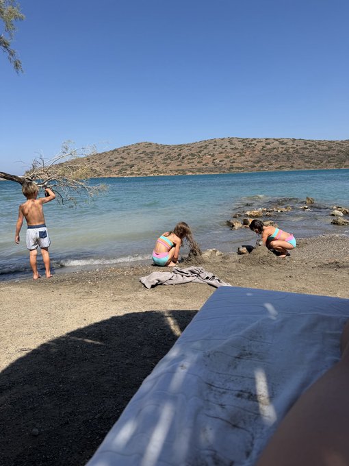 Three children play at the edge of a clear blue lake surrounded by hills, with a towel visible in the foreground on the sandy shore.
