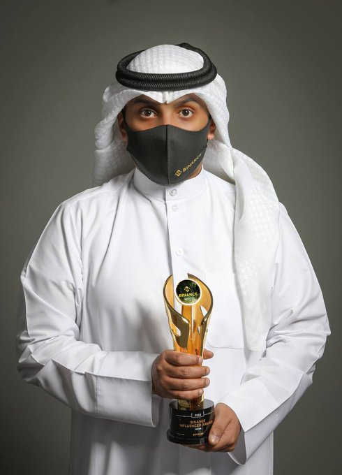A man wearing a traditional white thobe, keffiyeh, and a black Binance-branded mask, holding a golden Binance award trophy.