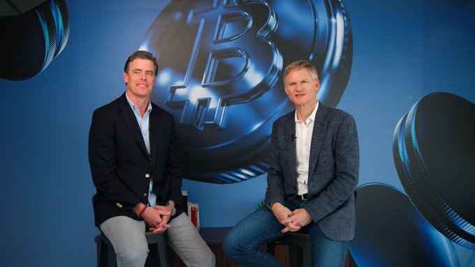 Two men in business-casual attire sit in front of a blue backdrop featuring a large 3D Bitcoin symbol.