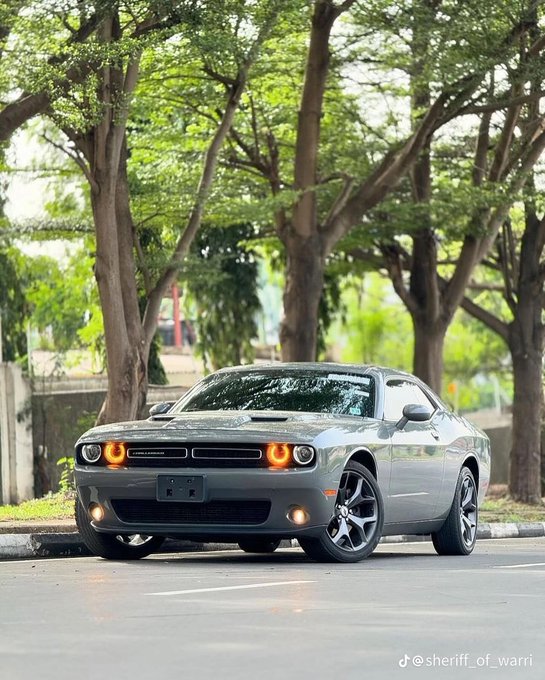 A photograph of a silver Dodge Challenger parked on a street, featuring distinctive halo headlights, with large trees and greenery in the background and a TikTok watermark '@sheriff_of_warri' at the bottom right.