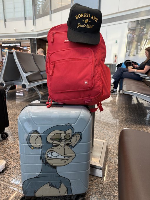 An airport waiting area with a red backpack topped by a 'Bored Ape Yacht Club' cap, resting on a suitcase featuring a cartoon monkey graphic.