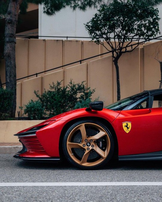 A red Ferrari sports car with gold alloy wheels and the iconic prancing horse logo parked on a street beside a beige wall with trees and shrubbery in the background.