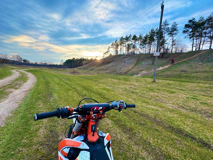 First-person view from an orange dirt bike on a grassy path at sunset.