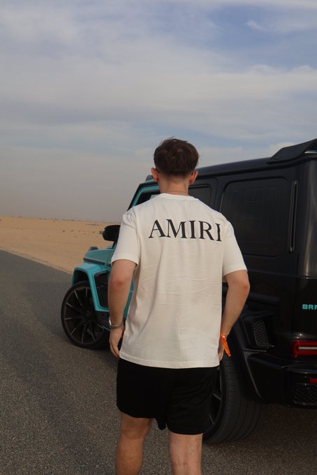 A man wearing a white "AMIRI" t-shirt and black shorts stands in front of a turquoise and black luxury SUV in a desert setting, embodying the "GM (get money)" lifestyle through designer fashion and high-end vehicles.