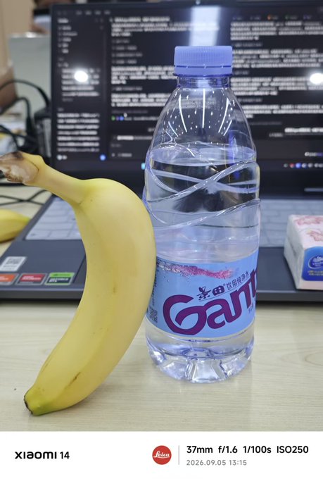 A still life photo taken on a Xiaomi 14 showing a banana leaning against a bottle of 'Ganten' water on a wooden desk in front of an open laptop displaying code.