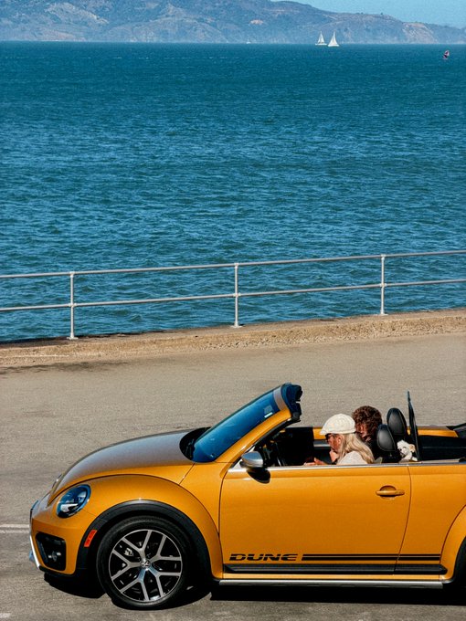 An orange Volkswagen Beetle Dune convertible with two passengers driving along a coastal road, with sailboats visible on the blue ocean and mountains in the background.