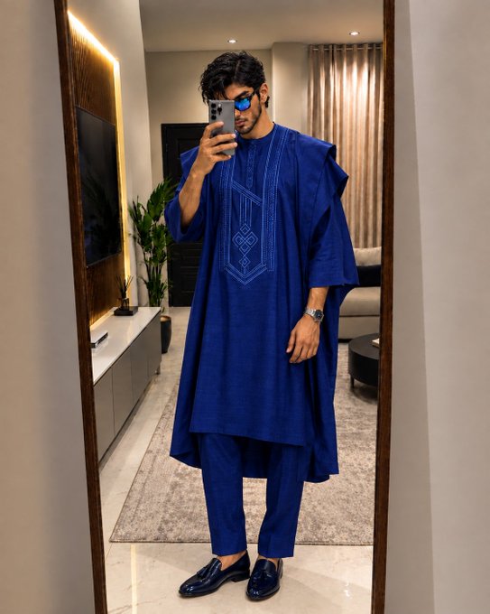 A man in sunglasses takes a mirror selfie wearing an elegant blue traditional African tunic and pants outfit with matching loafers.