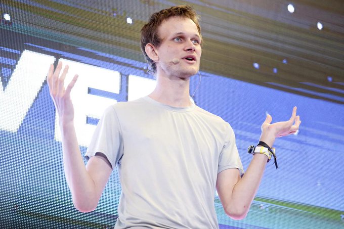 A young man with short brown hair, wearing a light gray t-shirt and a headset microphone, gestures expressively while speaking on stage against a digital backdrop displaying partial text. The image illustrates the article about Vitalik Buterin unveiling the top priorities in Ethereum's new "Lean Ethereum" roadmap, which focuses on quantum-resistant security, scalability, and privacy enhancements over the coming years.