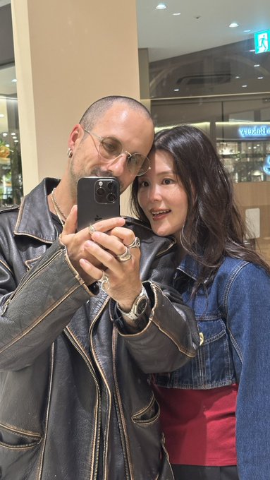 A mirror selfie of a couple in a shopping mall - a man with a shaved head wearing a black leather jacket and sunglasses holding a phone, and a woman in a denim jacket leaning beside him.