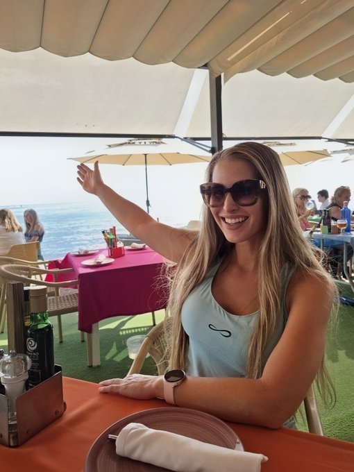 A smiling woman wearing sunglasses and a light blue top sits at an outdoor seaside restaurant table with a red tablecloth, waving her hand toward the ocean view.