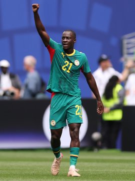 A Senegalese soccer player wearing the number 21 green and red jersey celebrates on the field with his fist raised.