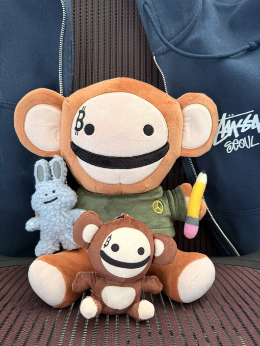 Two orange monkey plush toys with smiling faces, one larger and one smaller, sitting on a striped surface alongside a gray rabbit plush and a yellow crayon, with a blue Stüssy Soul jacket in the background.