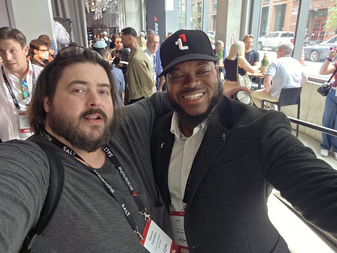 Two smiling men wearing lanyards pose for a selfie in a crowded convention hall, one wearing a black cap with a '1' logo.