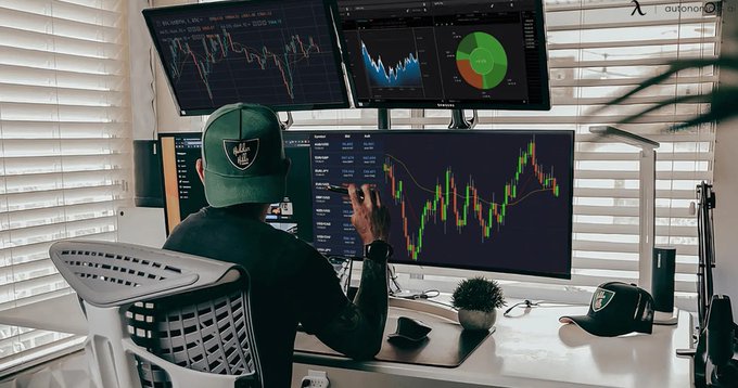 A person wearing a Raiders cap sits at a multi-monitor trading setup, analyzing candlestick charts and financial data.