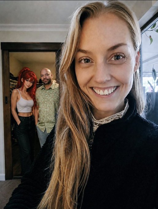 A selfie of a smiling young woman with long blonde hair wearing a black top, with a bald man in a green shirt and a woman with red hair in a white tank top standing in a doorway behind her.