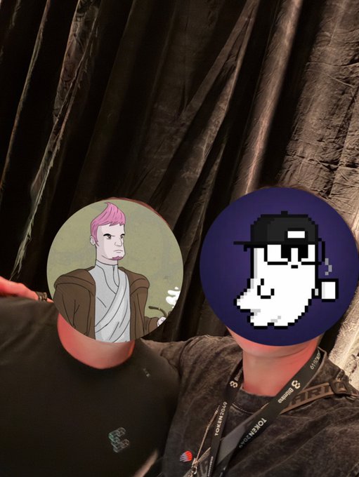 Two people are pictured with cartoon masks obscuring their faces; one mask shows a pink-haired character, and the other shows a pixel-art character smoking a cigarette. They are standing against a black curtain backdrop, with one wearing a Razer lanyard.
