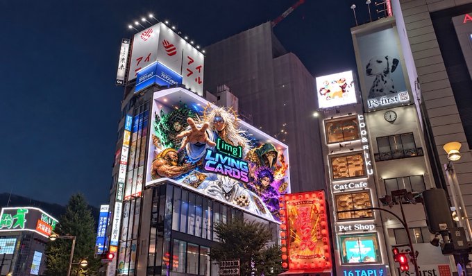 A nighttime cityscape, likely Tokyo, features brightly lit buildings, neon signs, and a large billboard advertising "img LIVING CARDS" above a "Cat Cafe" and "Pet Shop".