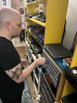 A photo of a bald man with a sun tattoo on his arm standing in what appears to be a music studio or shop, interacting with a wall of audio equipment and CDs.