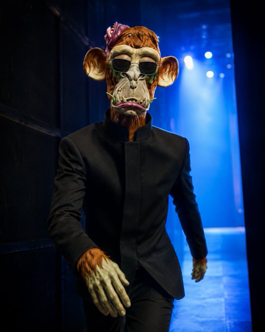 A person wearing a Bored Ape-style monkey mask with sunglasses and a black suit, walking through a blue-lit hallway.