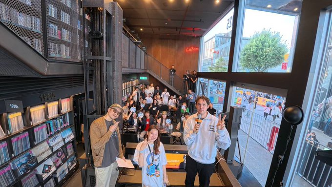 A lively indoor event takes place in a modern space with floor-to-ceiling bookshelves, where a crowd gathers while a man in a white sweatshirt and a young girl stand in the foreground near a staircase.