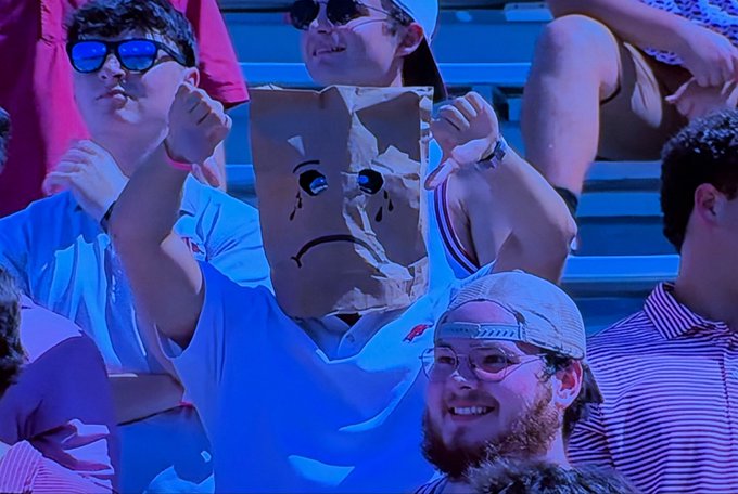 A person in a crowd wearing a white shirt holds up a brown paper bag with a sad face drawn on it, hiding their face.