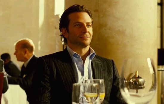 A cinematic shot of a man in a dark pinstripe suit and light blue shirt, smiling slightly in what appears to be a dimly lit, upscale setting with other figures in the background.