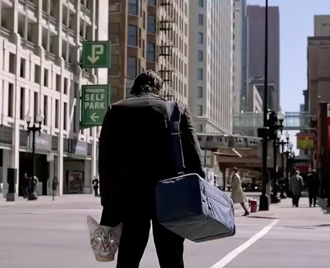 In the image, there's a man walking down a city street holding a cat mask over his shoulder while carrying a large suitcase. The caption of the image reads "The smallest feline is a masterpiece. Proton is the chosen one! #ProtonXPR". This suggests that the man might be participating in an event or promotion related to the movie "The Dark Knight" featuring the character Proton from the film.