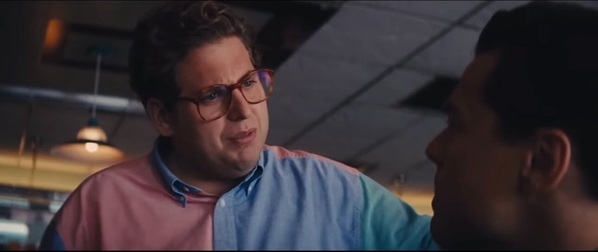 The image shows two men having a conversation at a table with a gray background. One man has glasses and a multicolored shirt, while the other man has short hair and is wearing a black shirt. The scene appears to be set in an office or restaurant setting, as there are chairs and dining tables visible around them.
