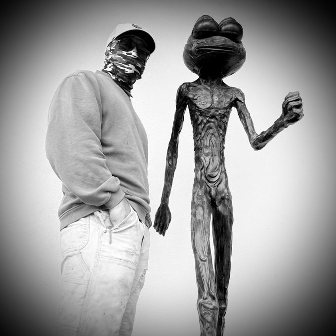 A black-and-white photo shows a person in a face mask and sweatshirt standing next to a glossy black frog-like sculpture.
