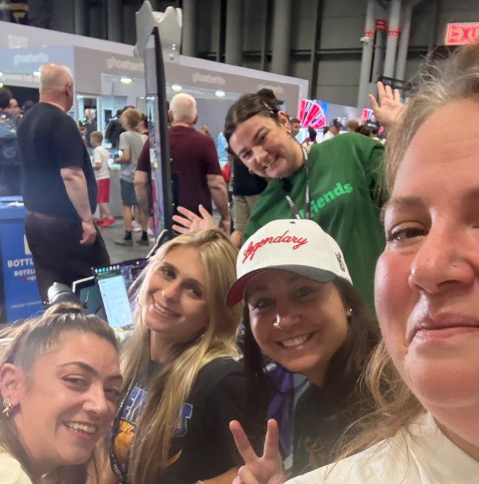 A group selfie of five smiling young women taken at a convention or expo, with one wearing a 'Legendary' cap and another in a green 'friends' shirt.