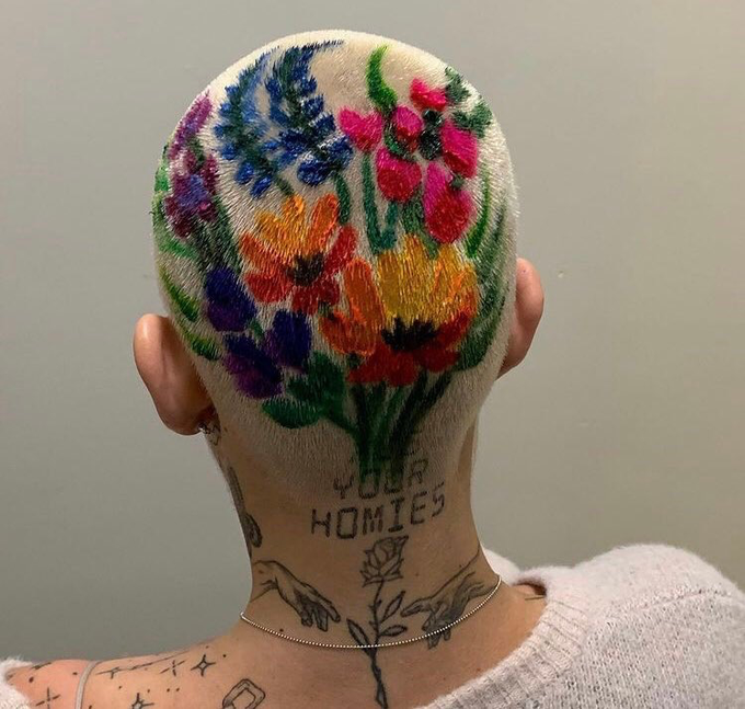 Close-up photo of the back of a person's shaved head featuring a colorful embroidered floral design with the text 'YOUR HOMIES' tattooed below, along with additional neck and shoulder tattoos.