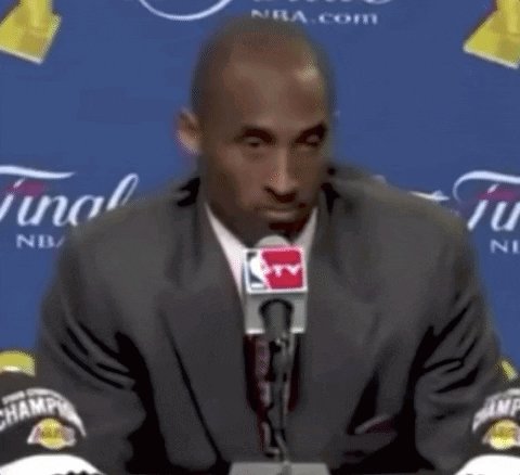 The image features a man wearing a suit and tie sitting at a microphone during an interview or press conference. The man appears to be a basketball player, possibly discussing his experiences or achievements with the media. The background of the image shows a blue wall adorned with yellow text, which could potentially be related to the event or organization being discussed by the man in the suit.