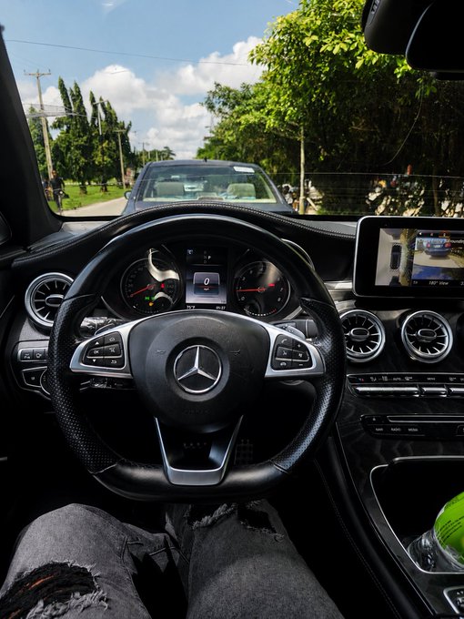 View from the driver's seat of a Mercedes, showing the steering wheel and dashboard with a road and tropical trees ahead.