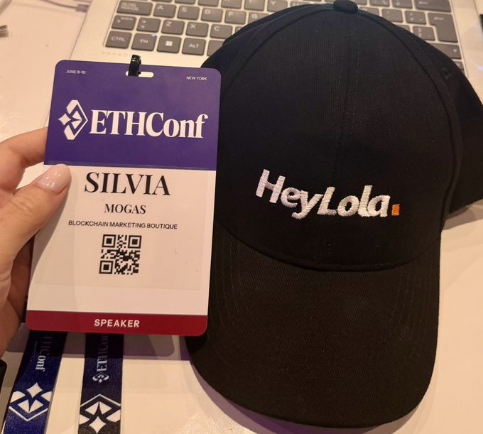 The image features a person holding up a black baseball cap with " HeyLola Dots Co. @ethconf" written on it, along with an electronic tag or lanyard that has a name tag attached to it. The person is wearing a pink nail polish and is holding the hat in their hand while looking at the tag.