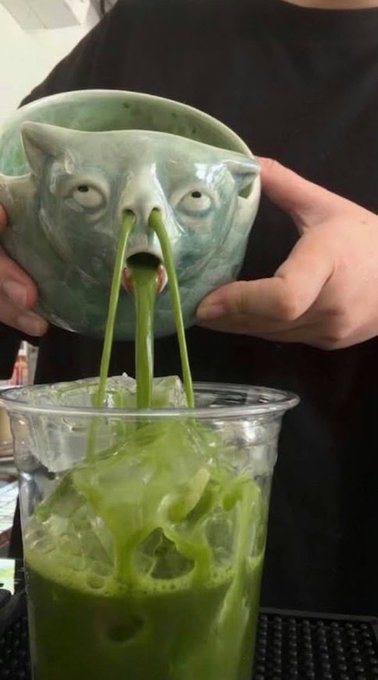 Hands hold a green ceramic bowl shaped like a cat's face, pouring a bright green liquid into a plastic cup filled with ice.