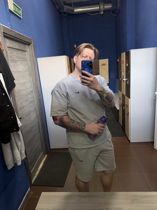 A person with blonde hair and tattooed arms takes a mirror selfie in a locker room, wearing a gray Nike t-shirt and shorts while holding a purple water bottle.