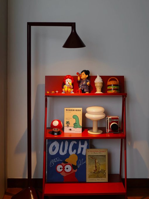 A red shelf with a brown floor lamp displays various items including a "1up" mushroom, a book with Chinese text, a "The Velveteen Rabbit" book, and a blue poster with the word "OUCH" featuring a cartoon bird.