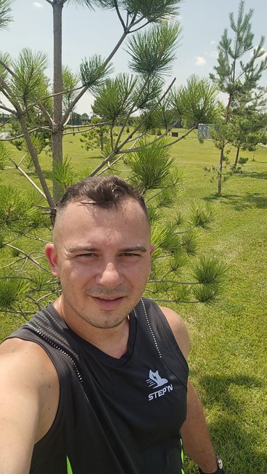 A man with a mohawk takes a selfie outdoors in a grassy park with pine trees, wearing a black athletic tank top that reads 'STEFN'.