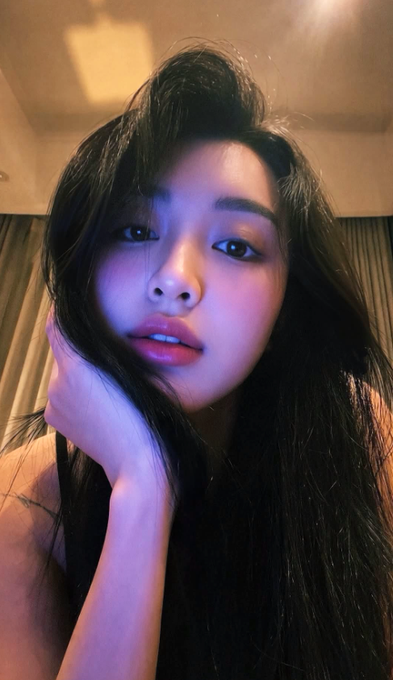 A selfie of a young woman with long dark hair, resting her face on her hand with a pensive expression, lit by purple and warm lighting.