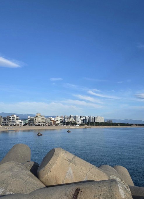 A coastal view looking across a body of water toward a city skyline and distant mountains under a clear blue sky, with large concrete tetrapods in the foreground.