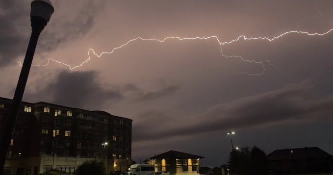 A nighttime photograph capturing a dramatic lightning bolt streaking across a dark, cloudy sky above silhouetted buildings and a street lamp.