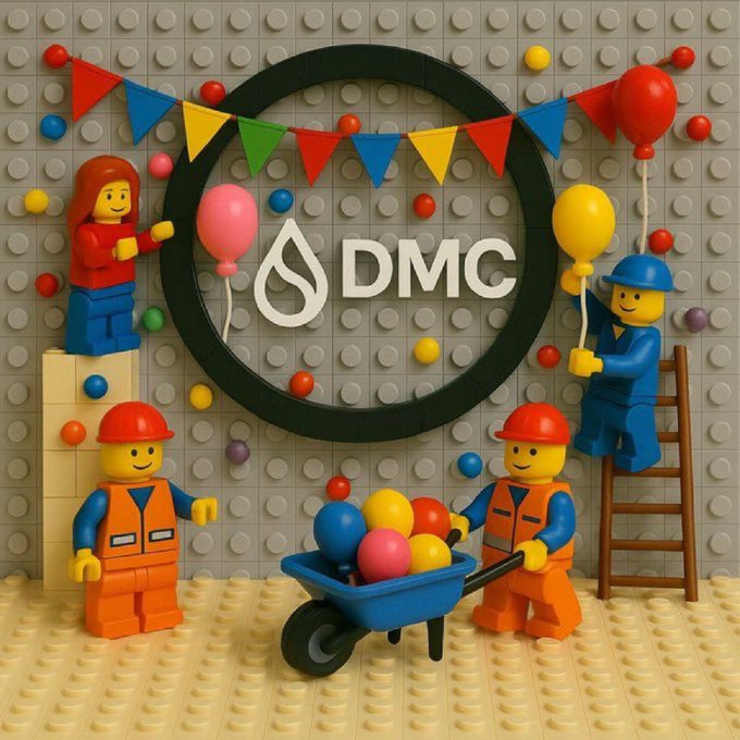 Lego construction workers celebrate around a circular "DMC" logo decorated with colorful balloons and bunting, with one figure pushing a wheelbarrow full of bright balls—visually symbolizing the steady, hands-on "consistent execution" the post credits for building long-term value around $DMC's blockchain and mobility vision.