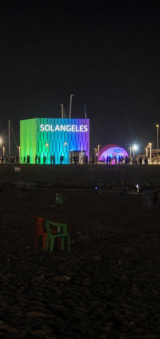 A nighttime beach scene showing a brightly lit building displaying 'SOLANGELE$S' in rainbow colors, with plastic chairs scattered on the sand in the foreground.